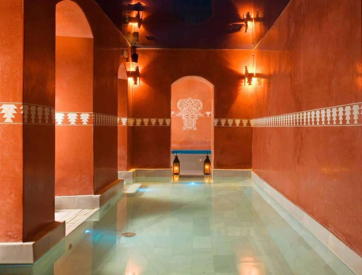 A long pool inside a hammam, surrounded by red walls with handing lanterns
