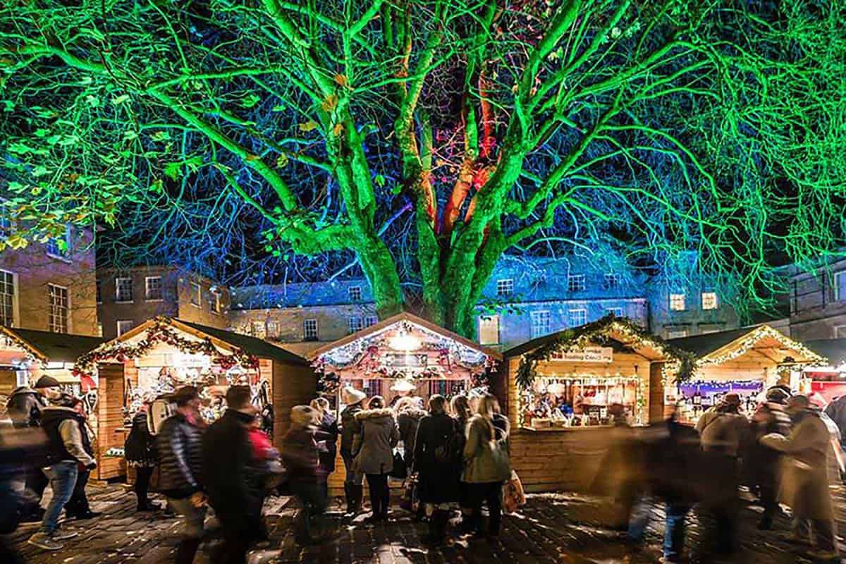Christmas market stalls with people, underneath a large tree lit up in green festive lights