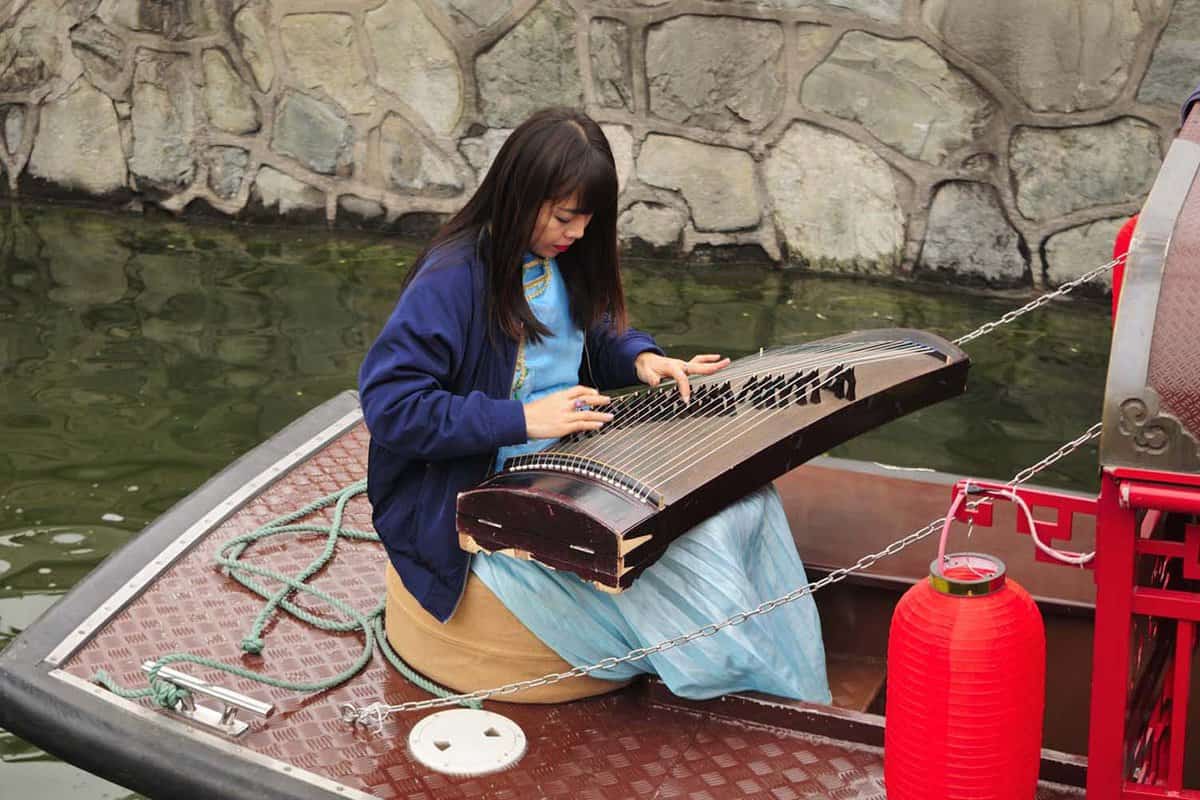 A woman sitting on a boat playing a traditional chinese instrument, guzheng, also known as the zither on the water canals near houhai bar street in Beijing China.