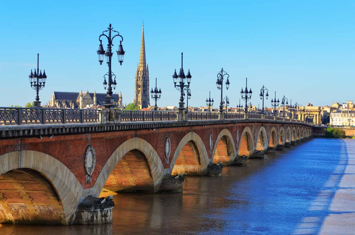 A red brick arched bridge with ornate lampposts crosses a wide river in Bordeaux