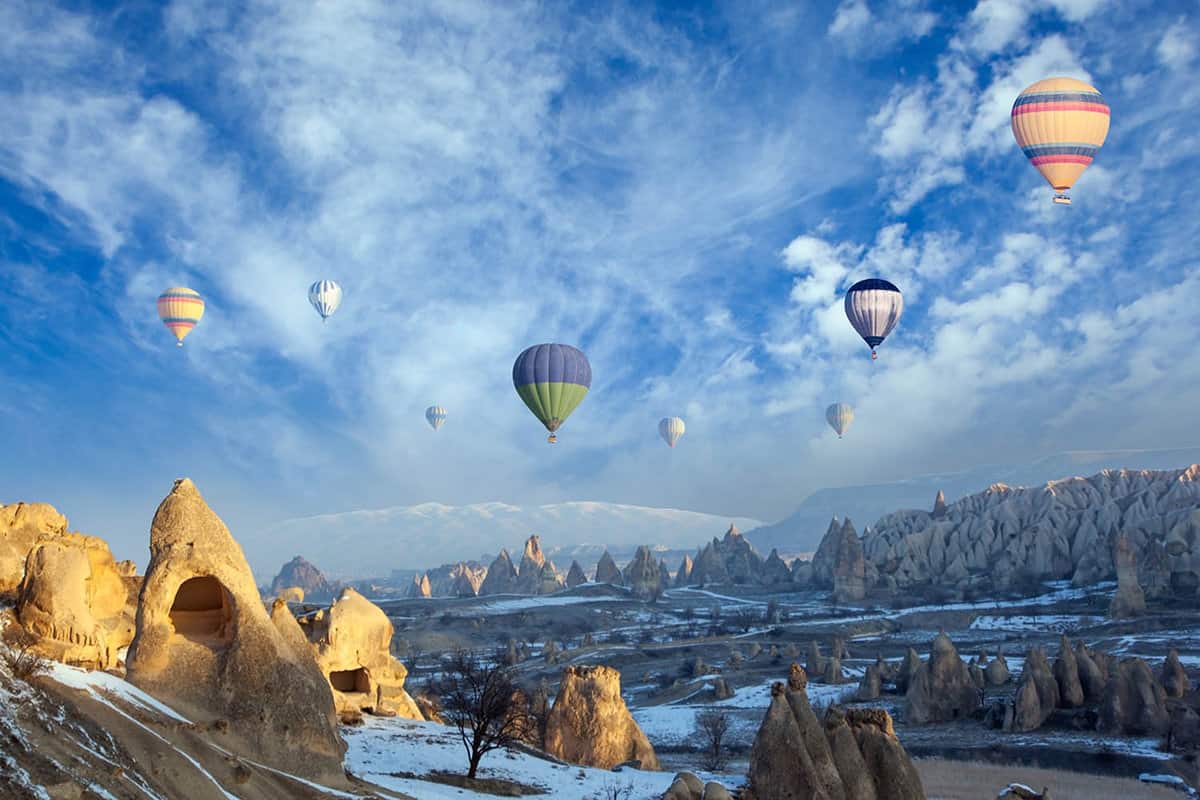 A view from the ground of many hot air balloons in the sky over a rocky landscape.