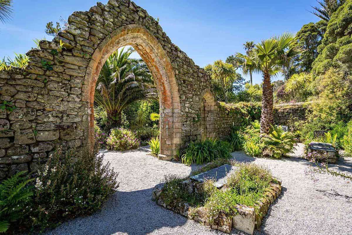 The ruined archway of the old Abbey on Tresco, Isles of Scilly, Cornwall. Tropical palm trees and vibrant flowers surround the ancient ruins.