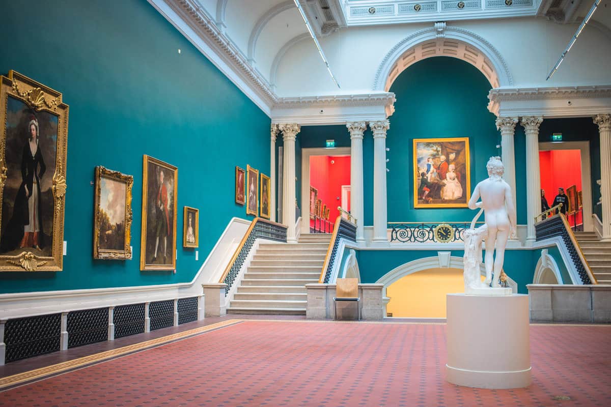 Inside a showroom of the gallery, with teal walls and white trim, oil paintings hanging on the wall, and a marble statue