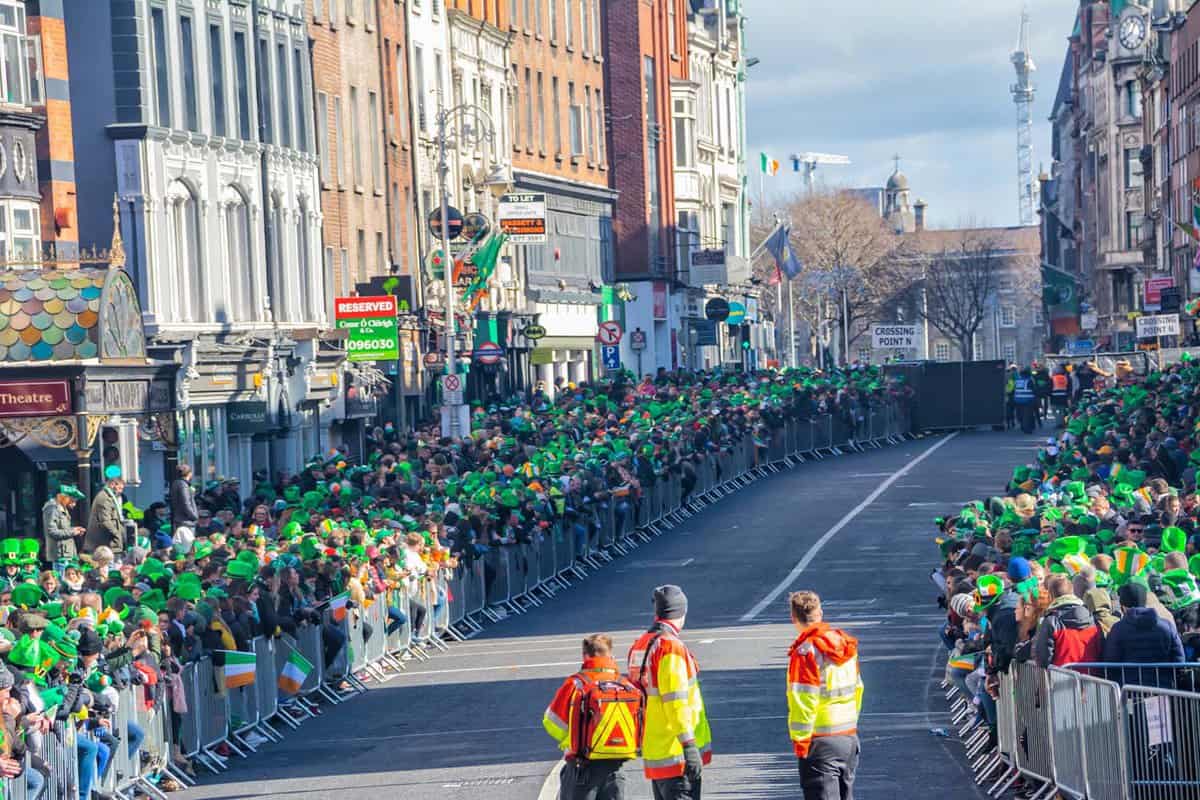Crowds of people wearing green hats with Irish flags stand on both sides of a street in Dublin waiting for the St Patrick's day parade.