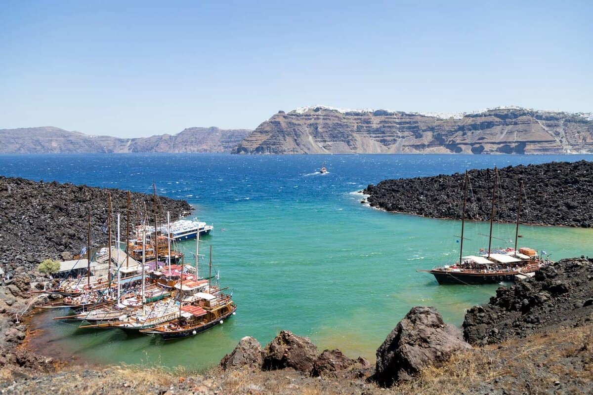 Sail boats sit in a blue and green harbor on the volcanic island Nea Kameni in Santorini, Greece