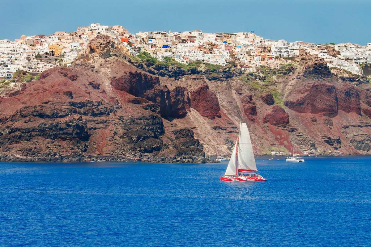 A sailboat sailing on bright blue waters in front of a cliff face in Santorini