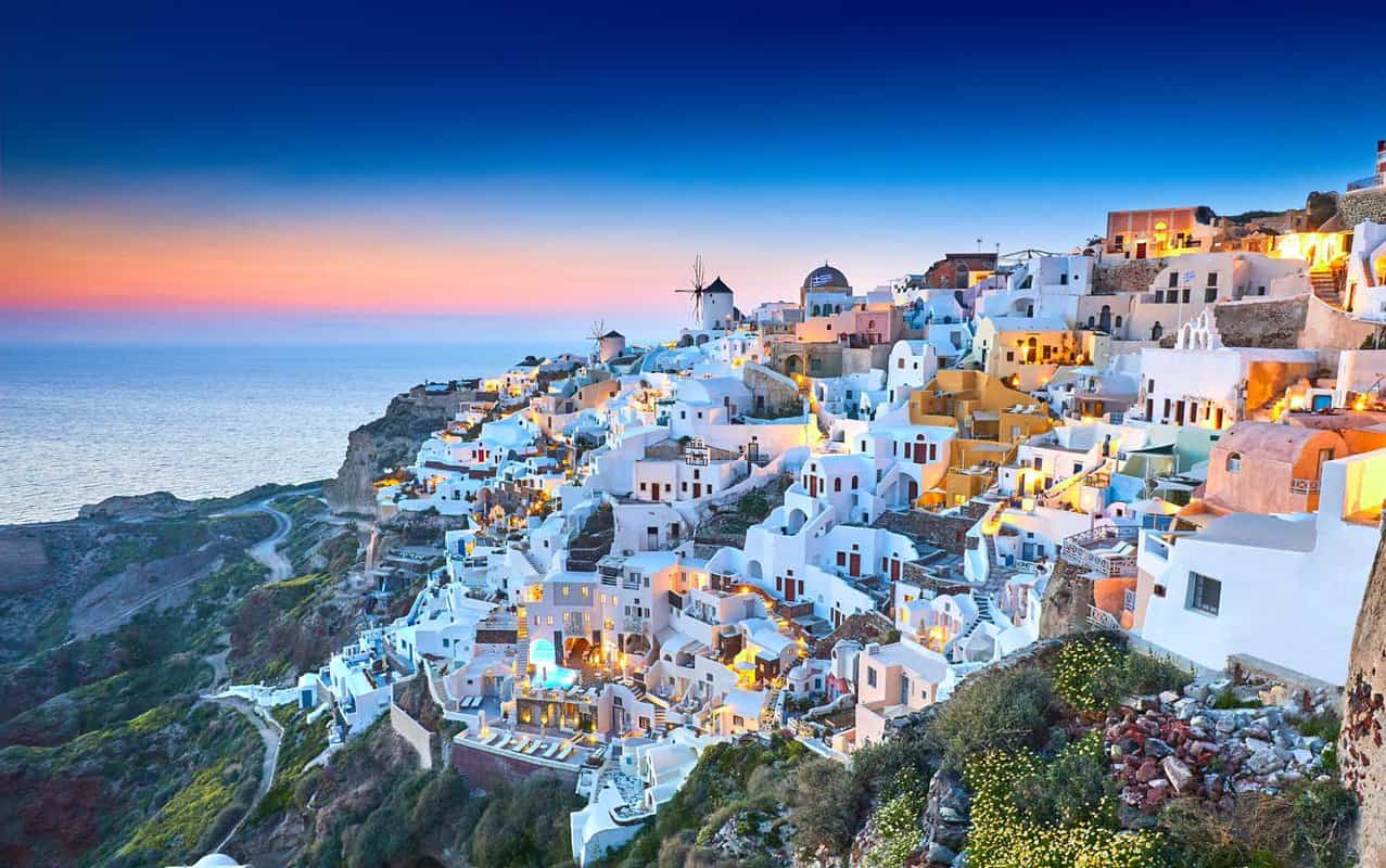 White Santorini buildings lit up in the early evening, with a sunset just beginning behind them