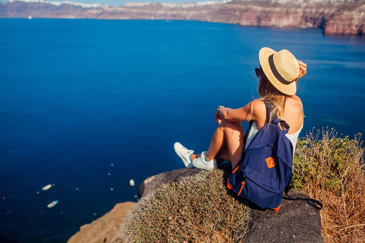 Woman wearing a sunhat sits on the edge of a rock and looks out over the caldera