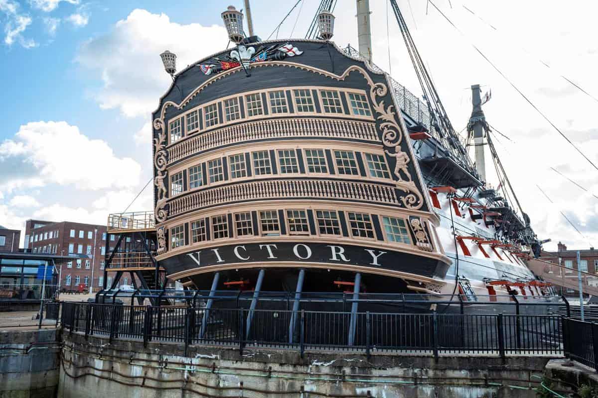 Stern view of HMS Victory, Lord nelson's flagship, on display at Portsmouth Dockyard. The ship is painted black with gold accents around the windows and the word 'Victory' is painted in white against the black exterior.