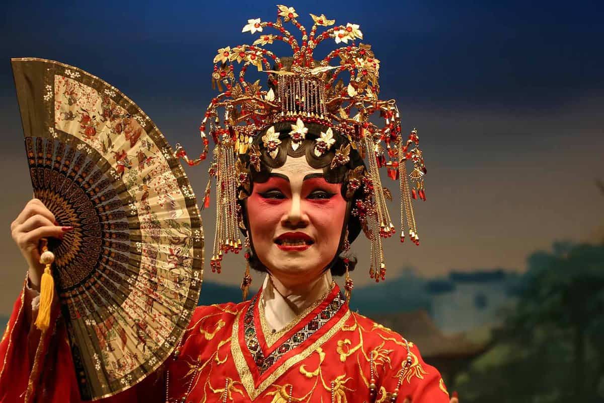 A Chinese opera singer in traditional wear, makeup and a golden headdress, holding a large fan to her face