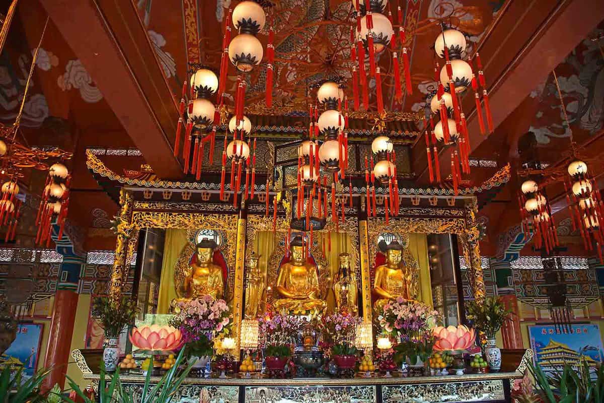 3 golden Buddhas inside the temple, surrounded by flowers and religious offerings