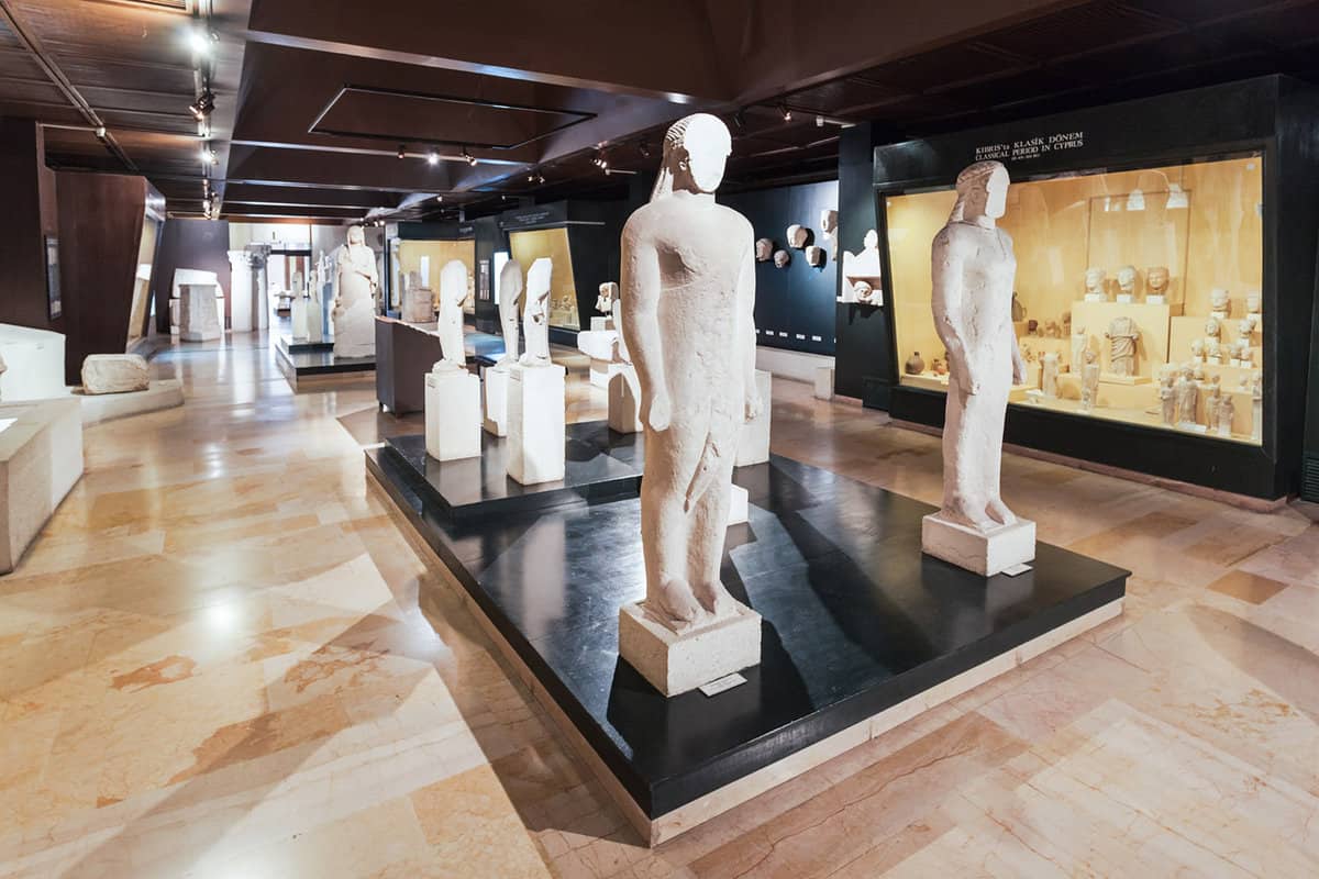 Faceless, white statues in a museum exhibit with many other statues and artefacts in the background