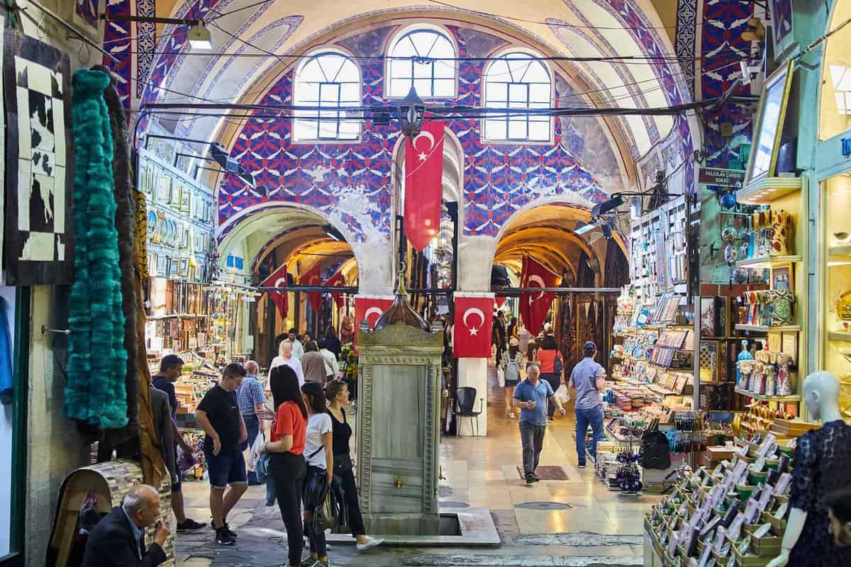 Interior of covered market with arched, tiled ceilings and Turkish flags hanging in the background as shoppers and shopkeepers wander through