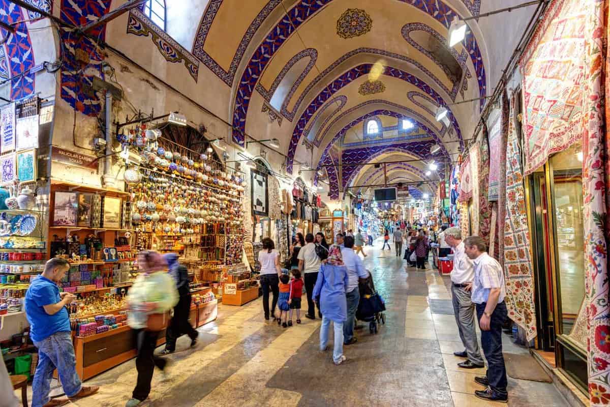 View down a long passageway in a covered marketplace with arched, tiled ceilings as shoppers pass by shopkeepers selling their wares