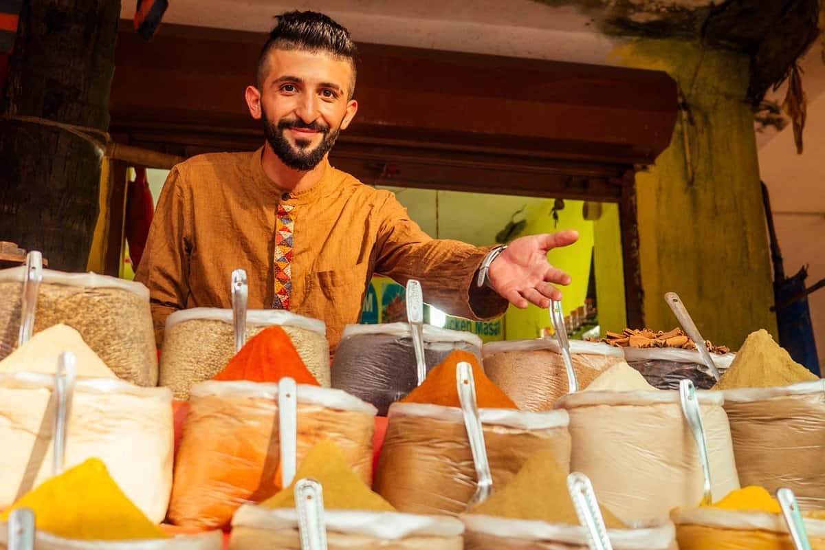 Young, bearded male gestures to triangular mounds of spices in his stall