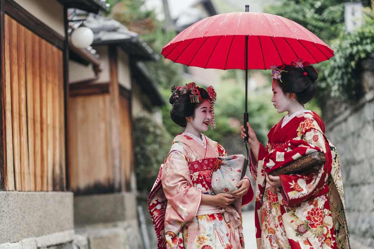 Two pink-clad geisha women talk beneath an umbrella in the street