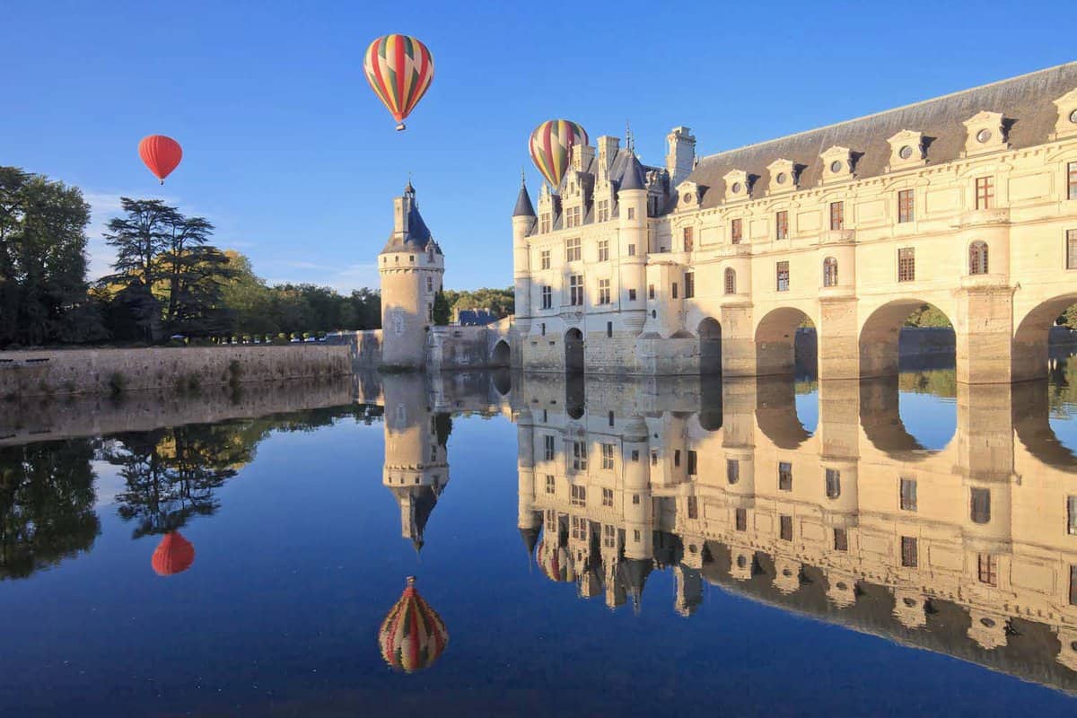 Three hot air balloons flying over Loire castle on a blue summer's day, reflecting on the river