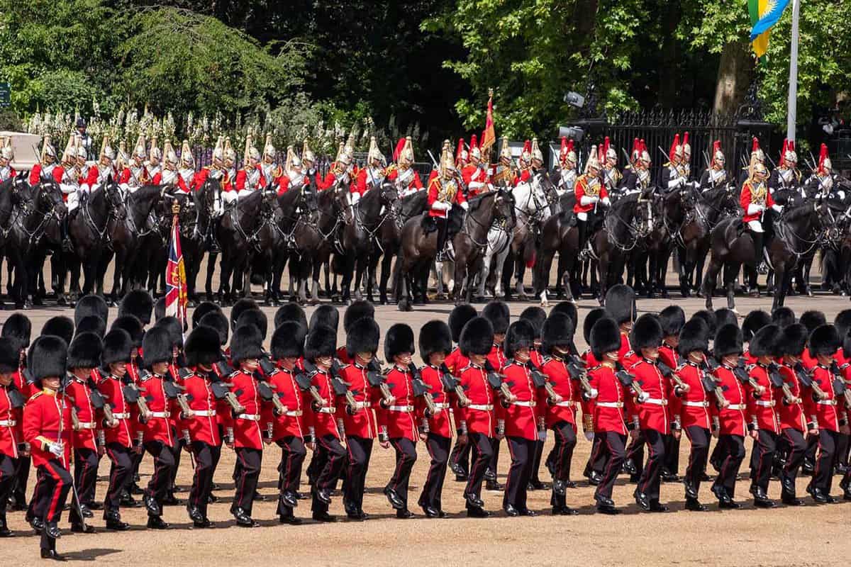 Close up view of soldiers in red uniforms and black hats parading, with horse guards behind