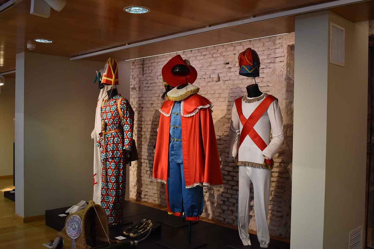 Spanish traditional costumes in a typical seventeenth century corrale. There are three costumes, the far left has patterned red and blue fabric and a red hat. The central costume has a gold-edged red cape, blue silky shirt and shorts, a floppy red hat, and a white ruff. The costume on the right has a white shirt and trousers. The shirt has a diagonal red X across the chest from shoulder to hip. A tartan hat is suspended above.