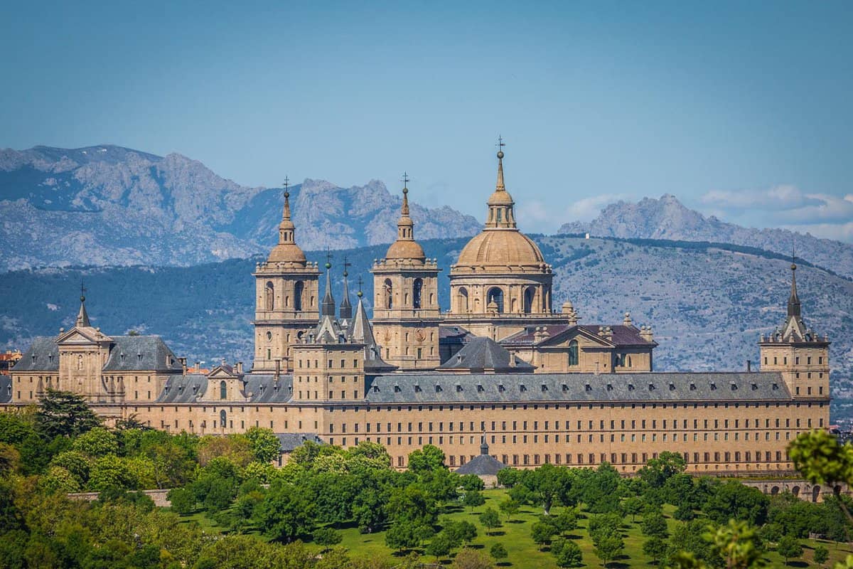 El Escorial Palace seen from a distance. Foliage sits in the foreground, while mountains rise in the background.