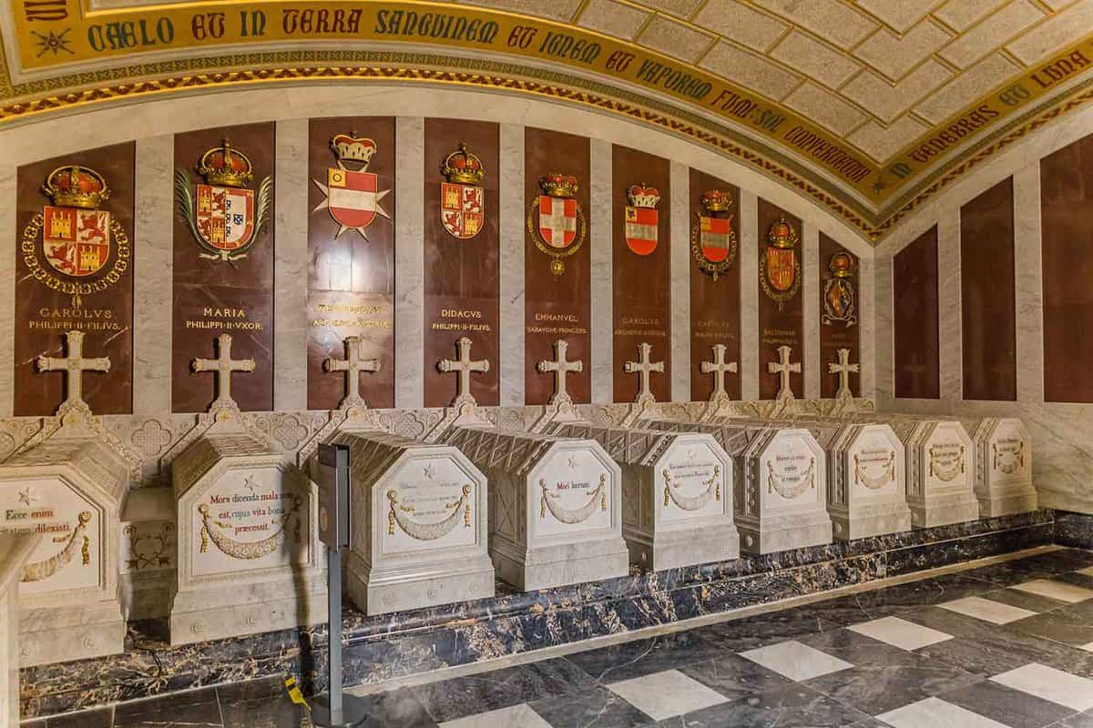 An image of the burial vault in El Escorial Palace. Stone sarcophagi line up in a row, raised from the ground by a dark grey marble plinth. Each sarcophagus is pale stone, with latticework etched along the top, and a banner with writing above it carved into the foot of the stone coffins (which is facing towards the viewer). Above each sarcophagus is a stone cross, and the wall above each coffin is dark red stone engraved with their name and decorated by their crest in full colour.