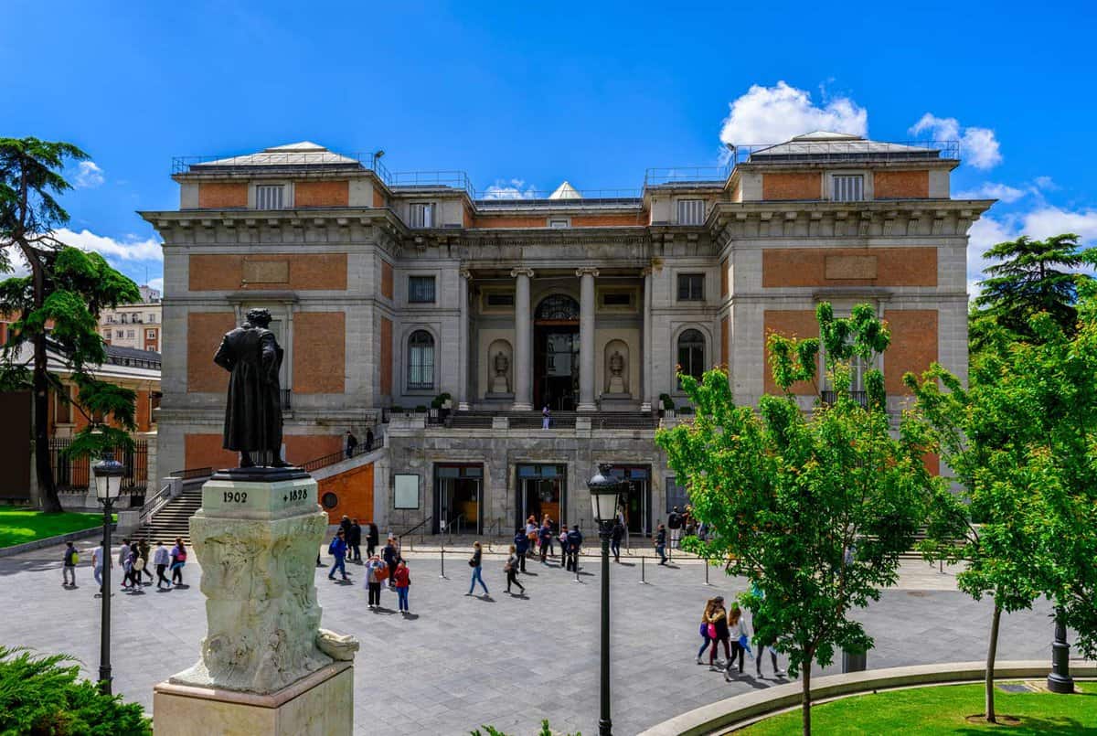 A statue of a figure sits in the left of the frame. In the centre is a plaza with people walking around, a few trees, and pale stone walkways. In the background is a Prado Museum building, made of orange and white brick.