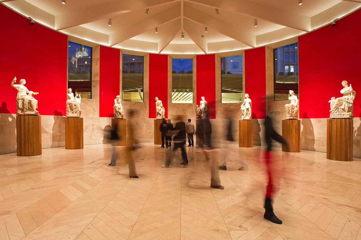 Marble statues stand on wooden plinths in a row, in front of red-painted walls. The windows leading outside show the dark blue of dusk. The image has been taken with a long exposure, and the blur of people walking around over time can be seen.