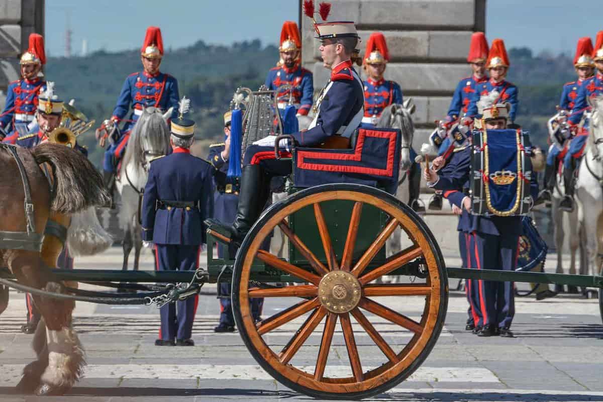 Guards during the Royal Palace of Madrid changing of the guards ceremony. A guard is pulled by a blue and gold painted horse and cart, and several guards sit on horses. Other guards stand in lines.