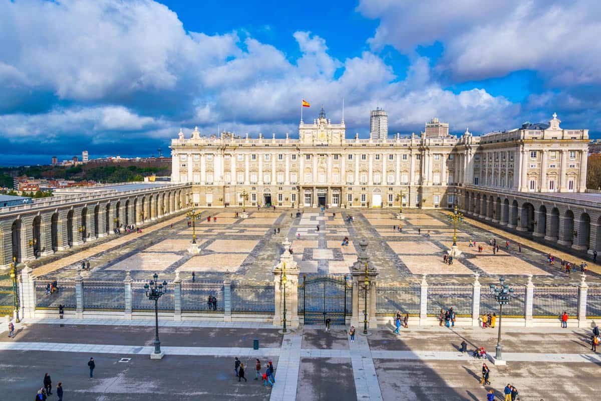 The Royal Palace of Madrid. The large, long, grand stone building has two walls running perpendicularly from it, and parallel to each other, on the left and right. The walls are made of dark grey stone and are a series of archways. There is a courtyard in front of the palace building, and a long wrought iron fence separating it from the public. You can see several tourists standing at the gates looking in.