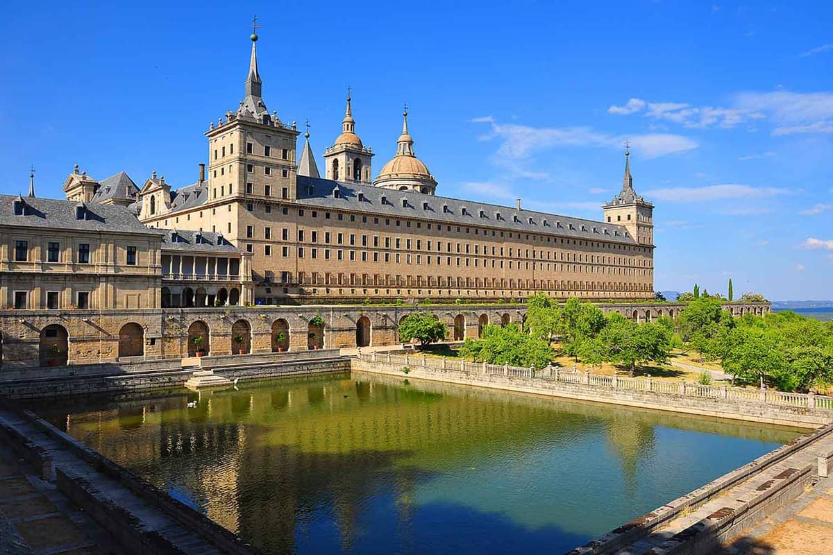 Exterior of El Escorial Palace in the late afternoon. A grand sandstone building with alot of small windows and a green-looking square pond in foreground