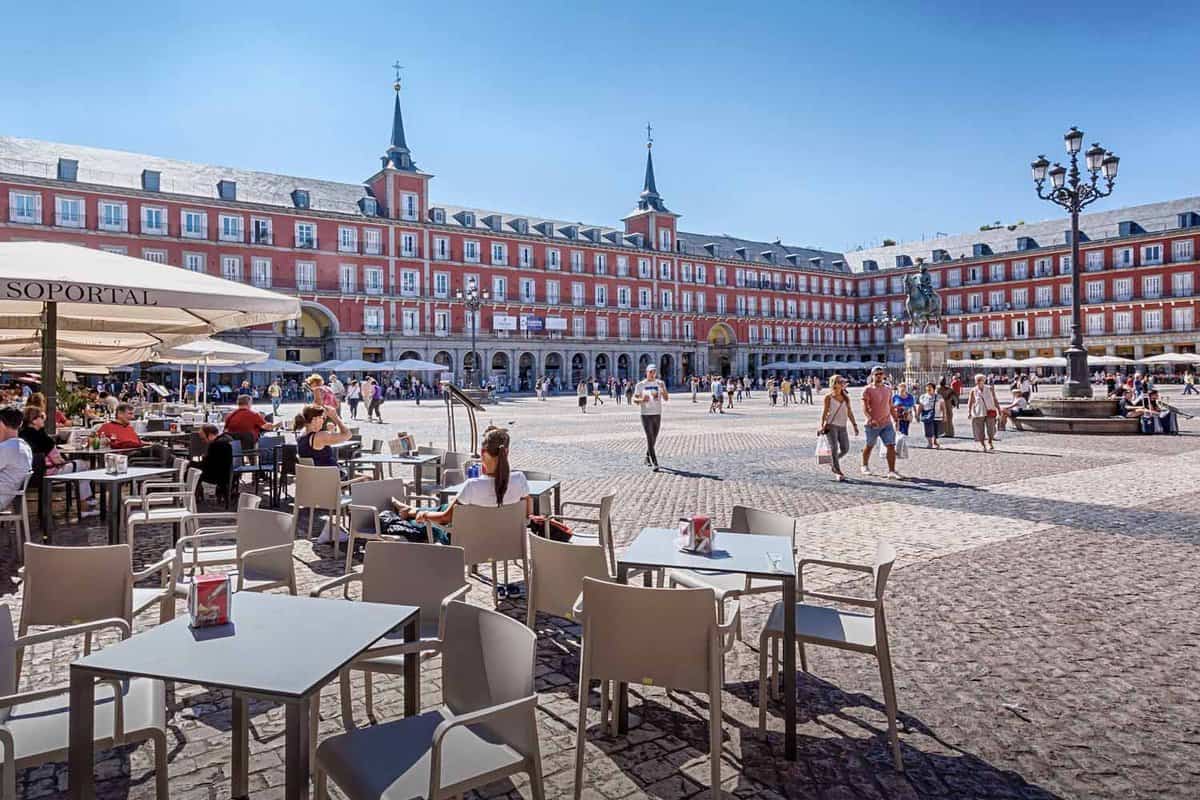 A view from the side of Plaza Mayor. Tables and chairs from cafes sit the the sun, people walk around on the cobblestone plaza.