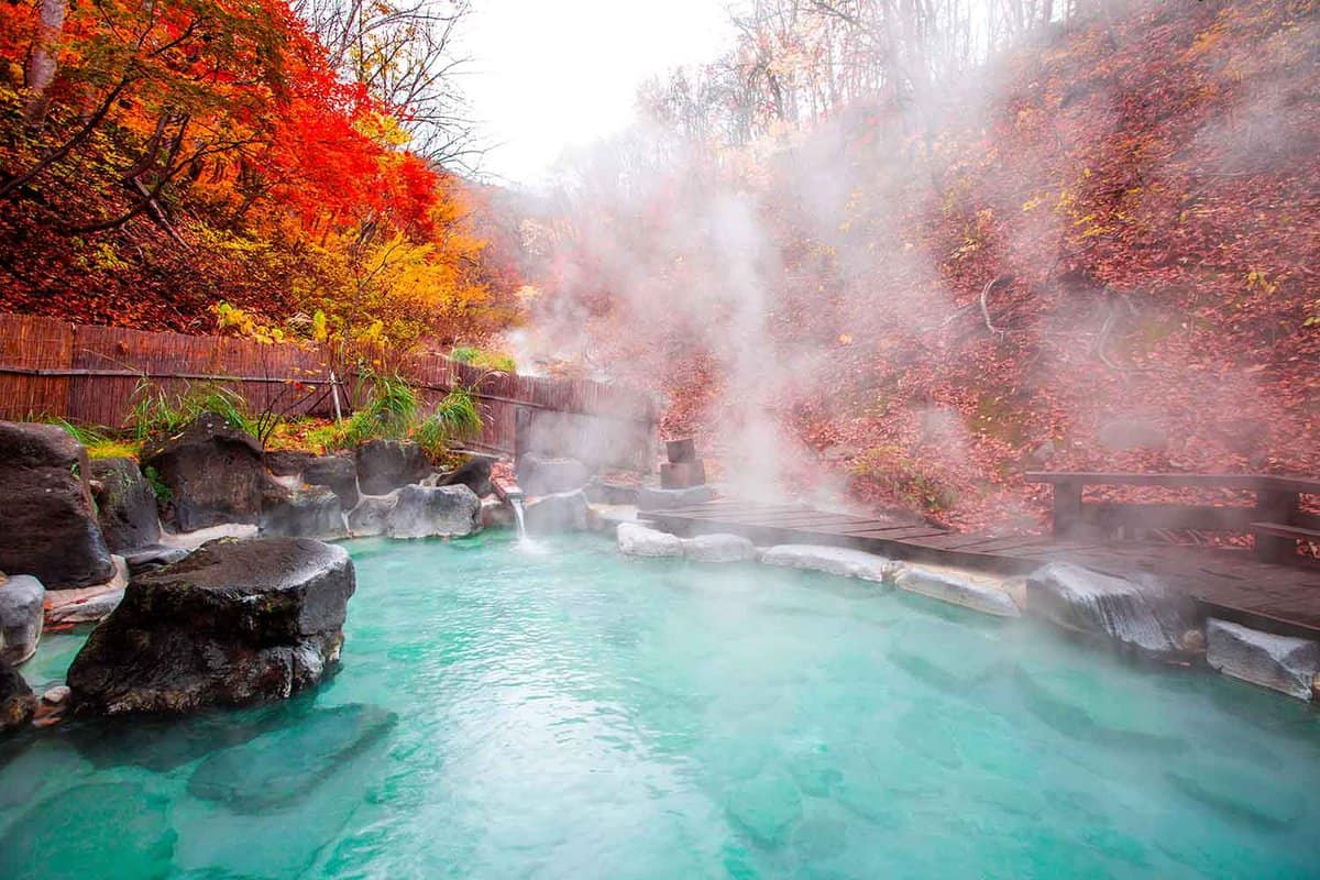 a large steaming outdoor onsen, surrounded by red-yellow leaves of acer trees