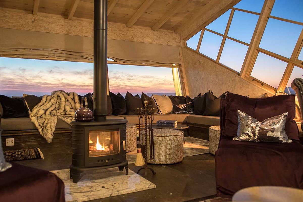 A log burning fire inside a lodge with views out at sunset