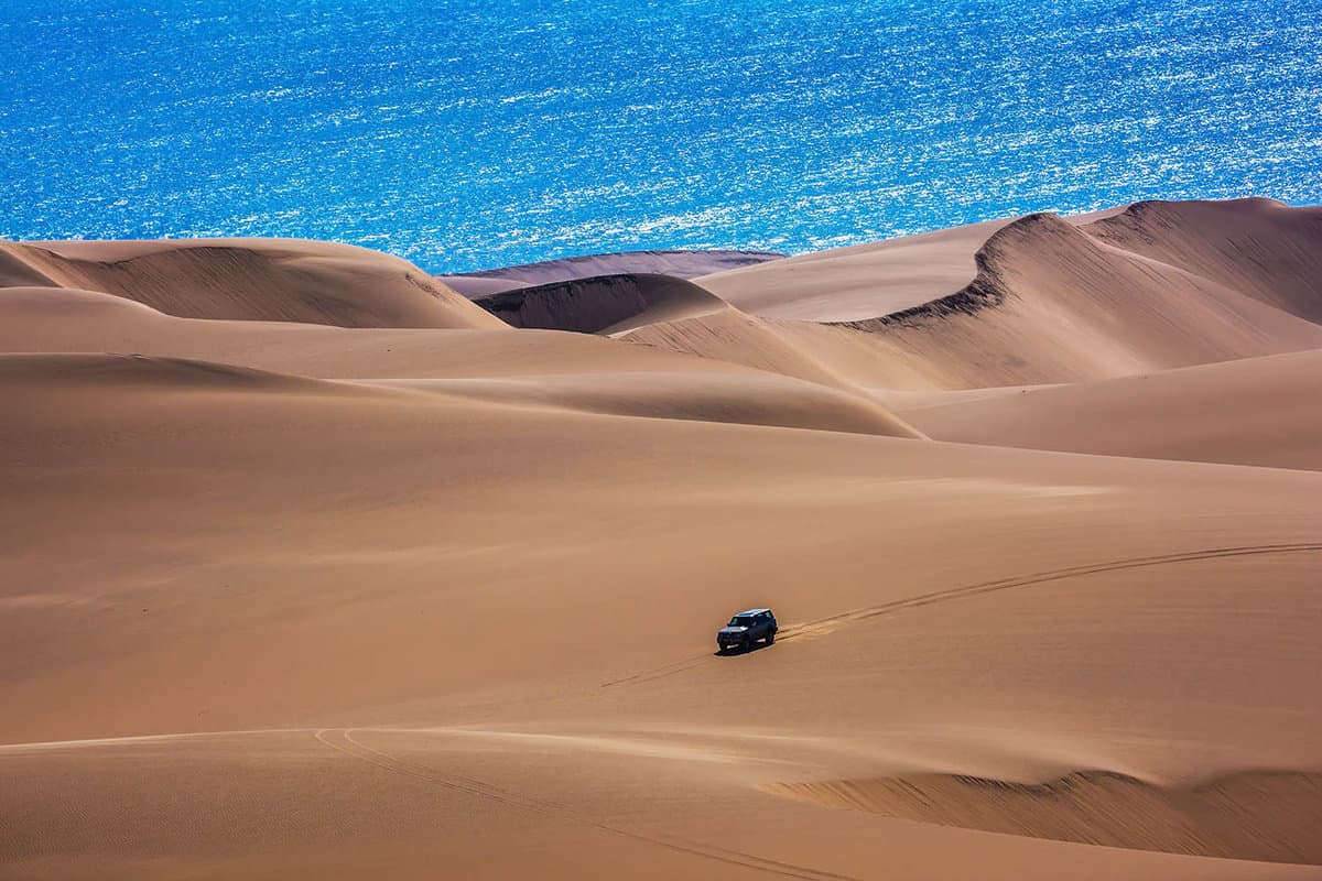 Sand dunes of the Swakopmund desert with a man in a vehicle and the blue sea in the distance