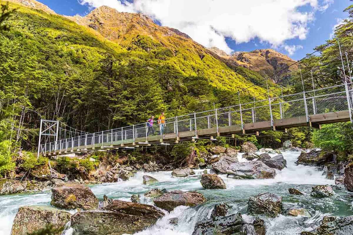 New Zealand tramping people crossing river bridge. Hikers couple backpackers walking hiking together with backpacks on Routeburn Track trail path.