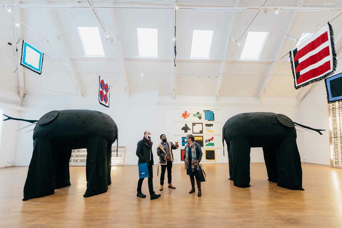 Inside the gallery. The room has pale wood floors, and high ceilings with skylights. There are people walking around large sculptures.