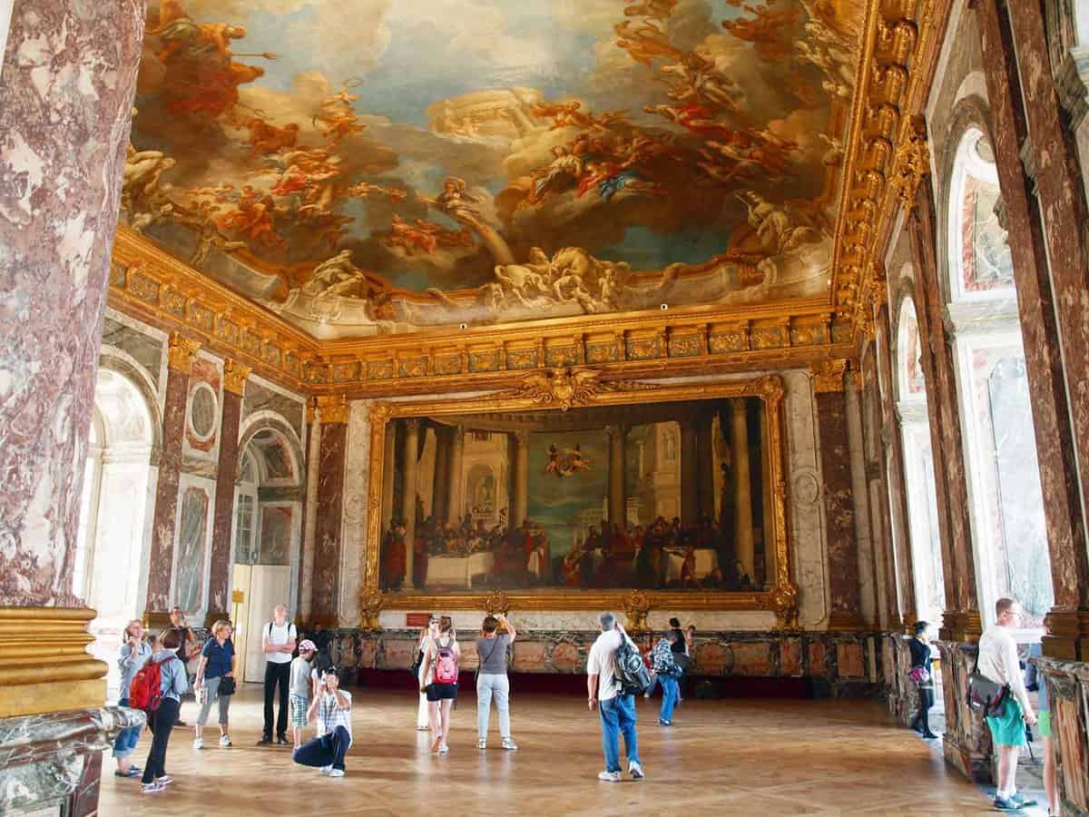 Inside view of a hall where there are tourists looking at huge gold framed paintings covering the front wall and a painted ceiling of 18th century art