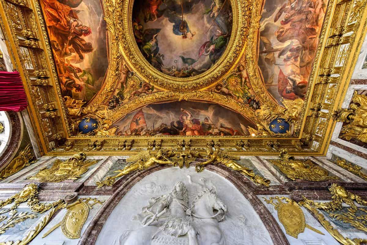 Looking upwards at the artwork of a wall and ceiling. The wall has engraved royal artwork with painted in gold frames. The entire ceiling above is painted with imagery of a battle with gold framing separate the different paintings.