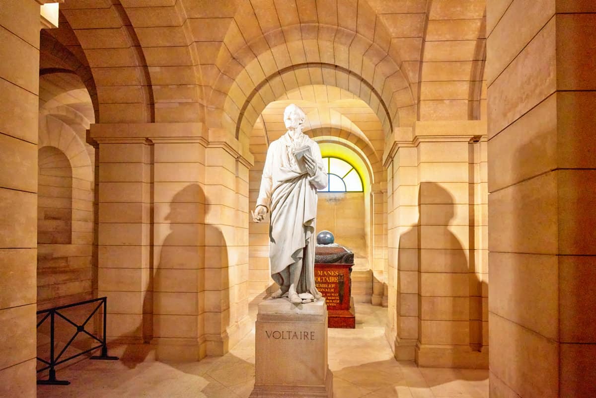 A statue and tomb of Voltaire in the Panthéon with a spot light on the statue creating two shadows