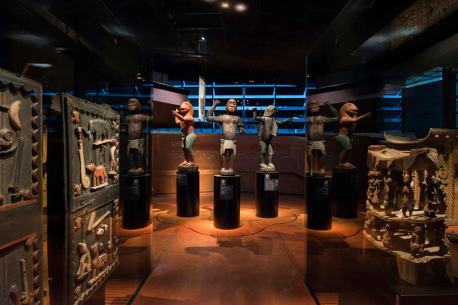 3 large carved wooden African-looking statues displayed on pedestals in a dark room