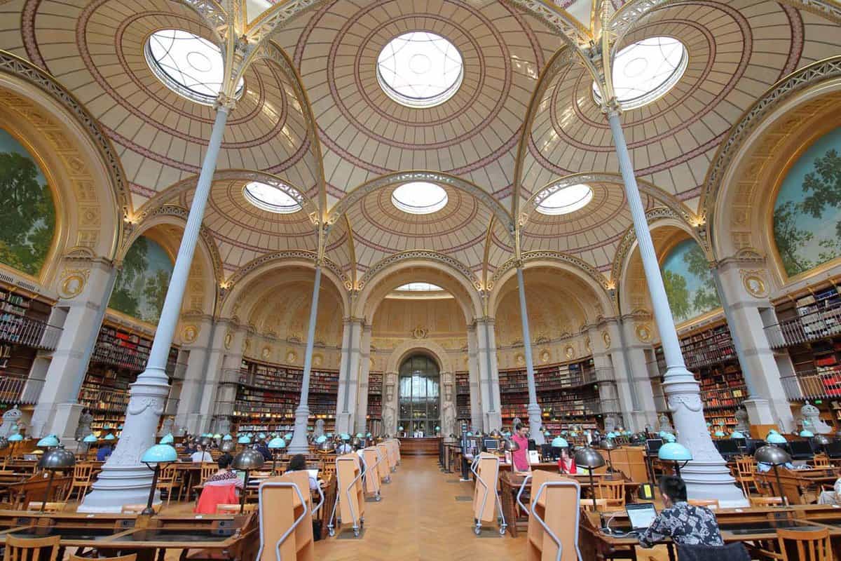 Unidentified people visiting the National Library of France. There are tall skinny podiums reaching the high ceilings, where there are round circular windows in the ceiling.