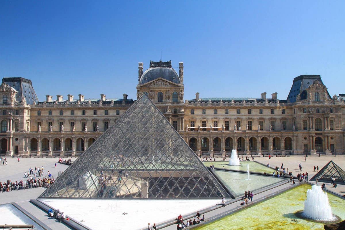 Landscape view of Louvre Museum outside building with the famous glass pyramid structure against a sunny blue day