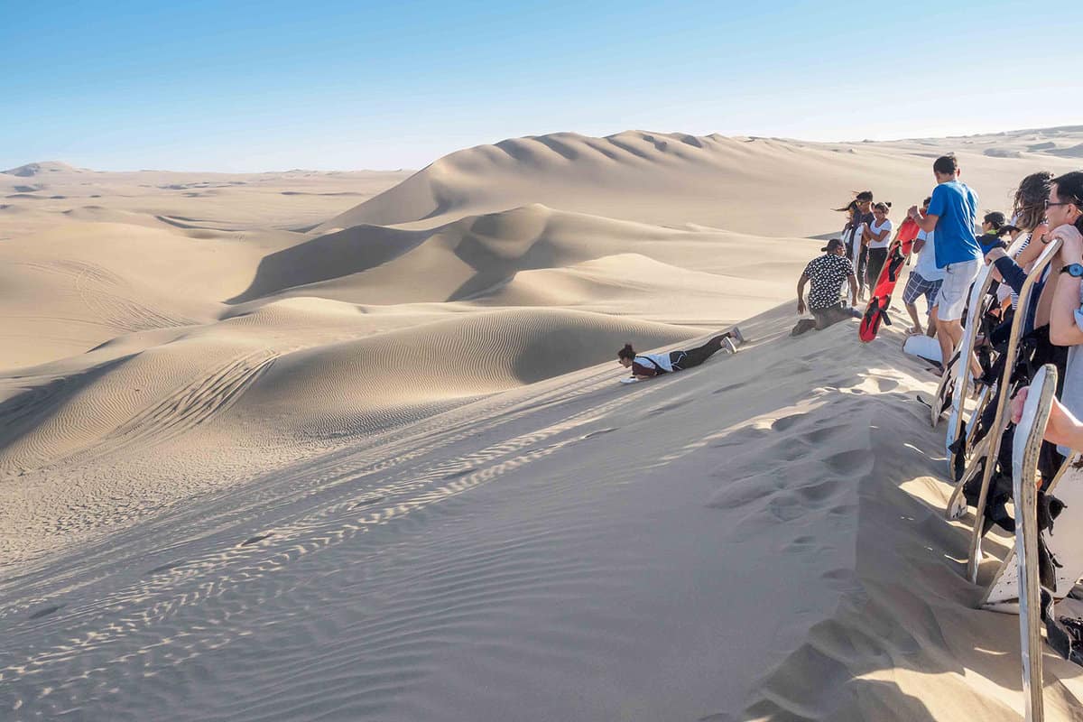Tourists stand on the edge of a sand dune ready to to sandboard