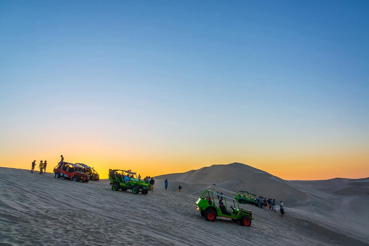 Tourists and sand buggies on a sand dune, with sunset behind them