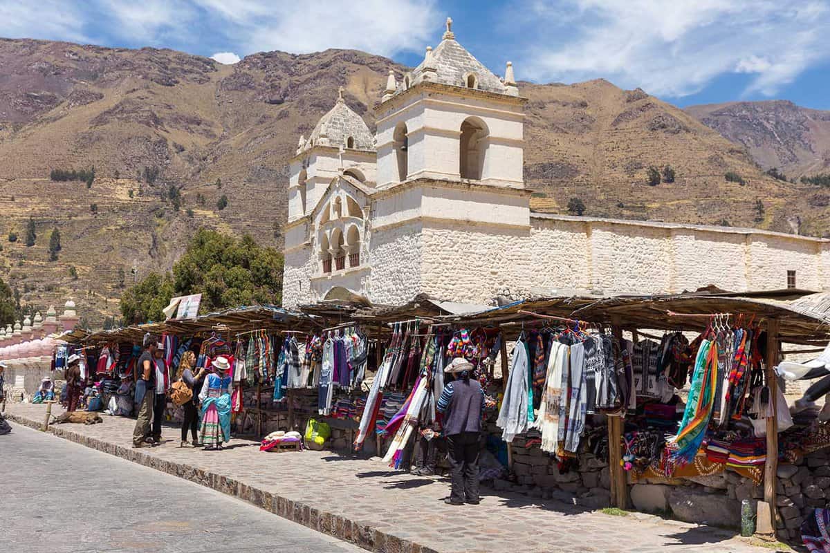 Stalls selling clothes and souvenirs line up in front of a white-brick church, with mountains in the background