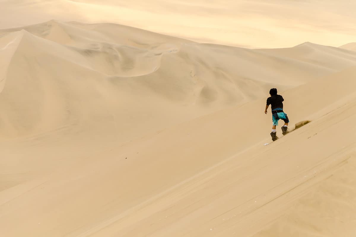 A person standing up and sandboarding down a large dune in Huacachina