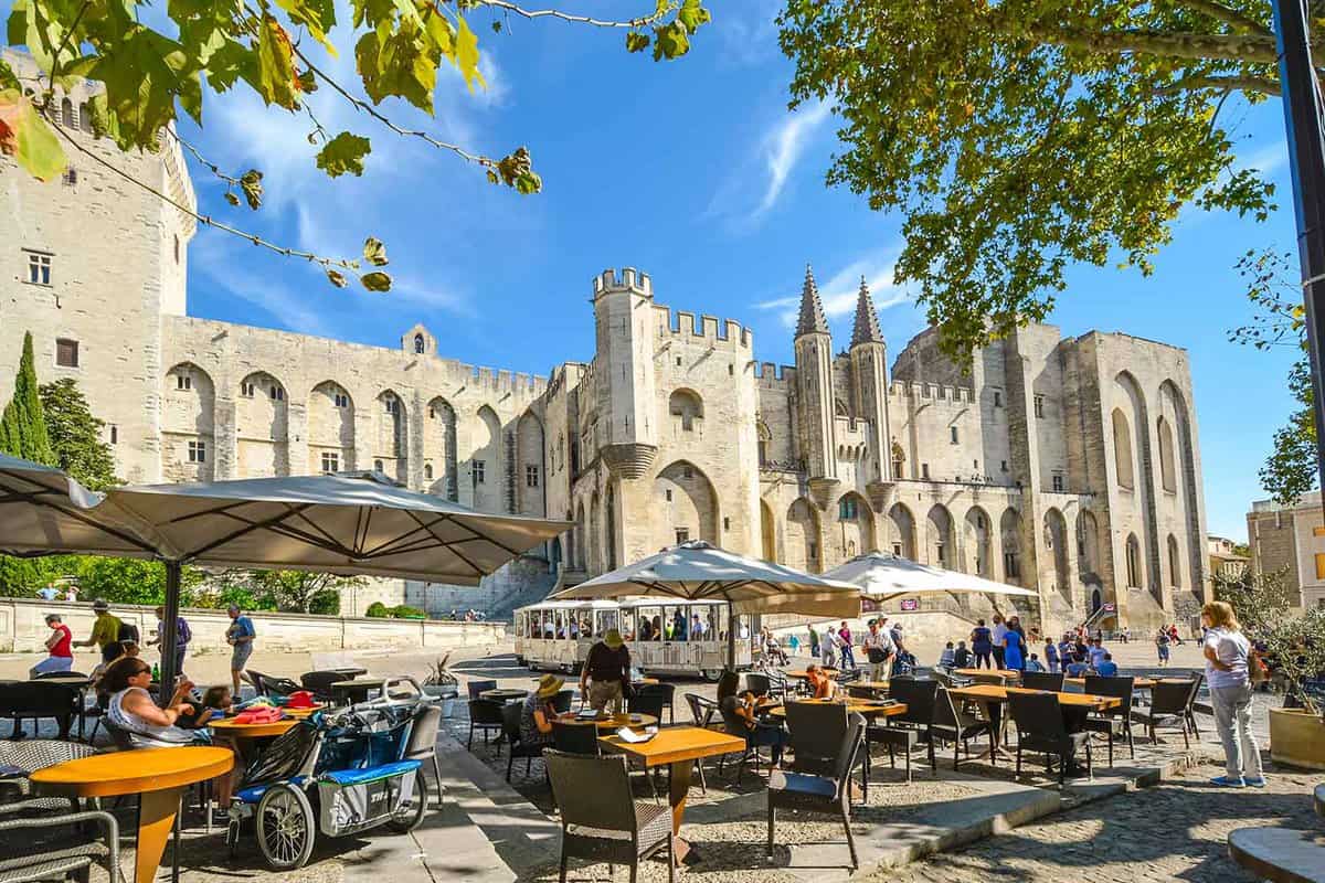 Tourists sit at a cafe in front of the medieval Pope's Palace while a tourist tram drives by on a sunny day