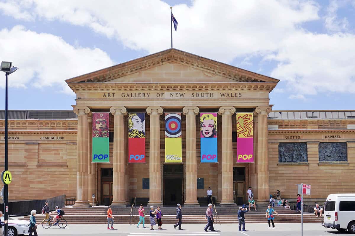 A grand building with 6 columns and colourful flags in between. People walking in the foreground.