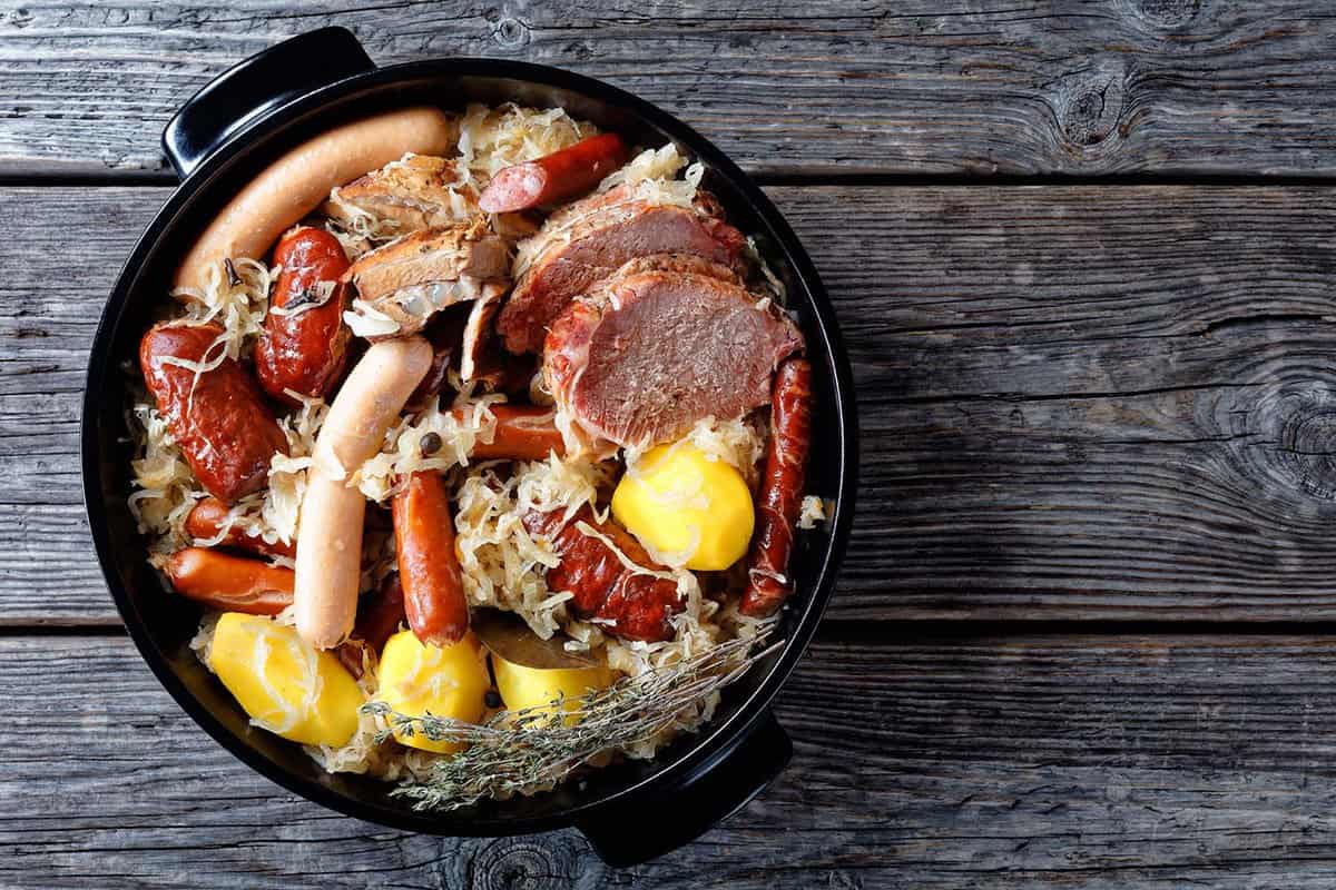 French choucroute garnie of sour cabbage with smoked bacon, pork loin, sausages potato and thyme stewed in white wine with onion, garlic served on a black baking dish on wooden background, top view