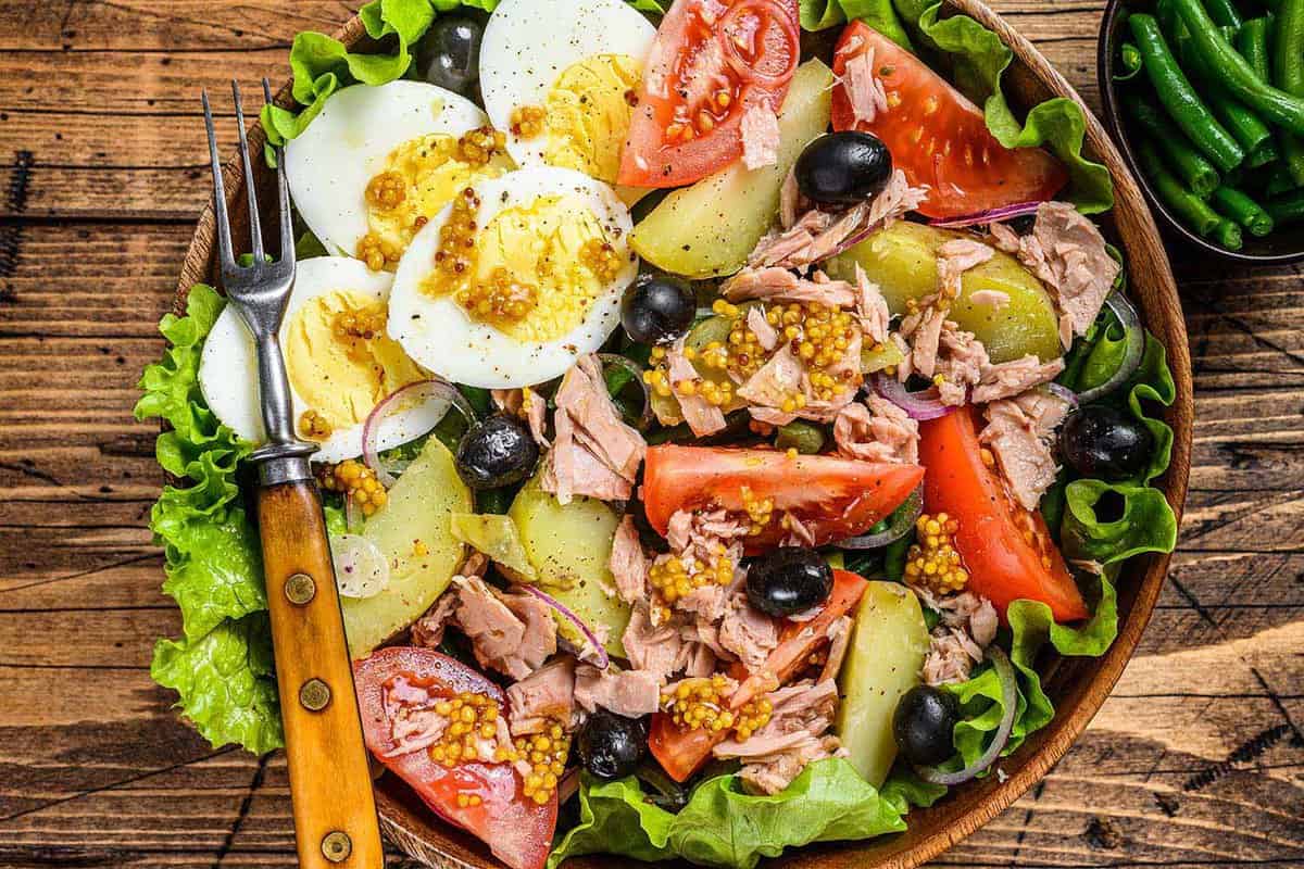 Nicoise salad with tuna, tomatoes, olives, green beans, cucumber, soft boiled eggs and potato in a wooden bowl. wooden background. top view.